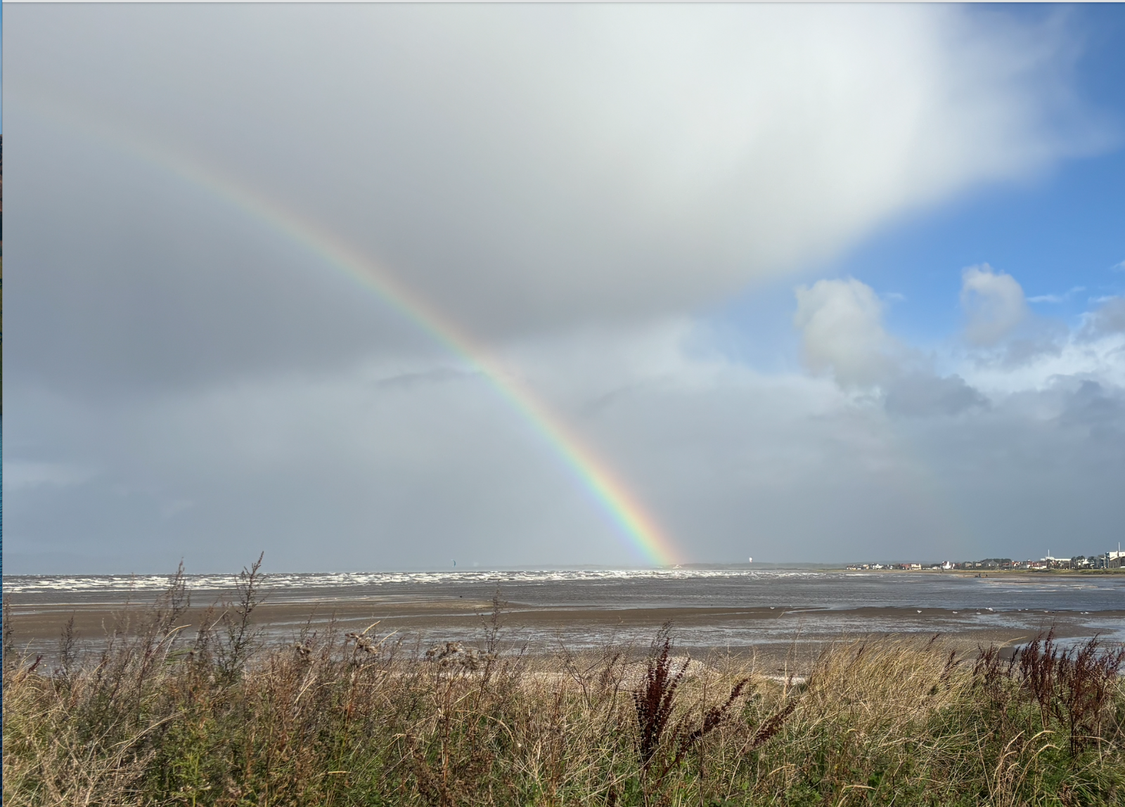 Troon coastline. Rainbow in cloudy sky over choppy waves, seen behind course grass and bushes.