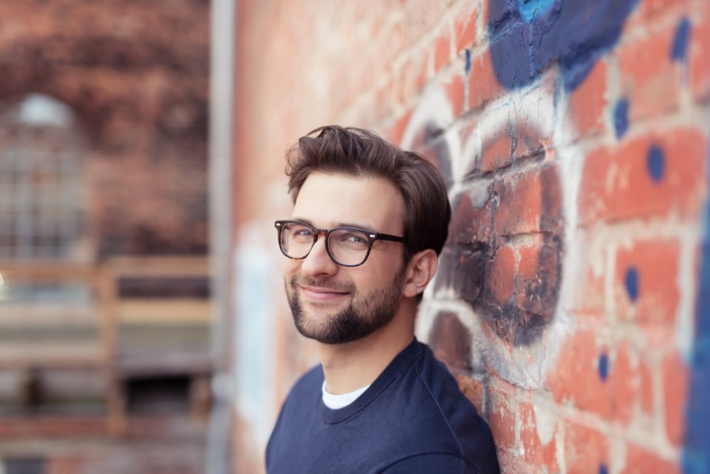 Portrait of Smiling Young Man with Facial Hair Wearing Eyeglasses and Leaning Against Brick Wall Painted with Graffiti Portrait of Smiling Young Man with Facial Hair Wearing Eyeglasses and Leaning Against Brick Wall Painted with Graffiti