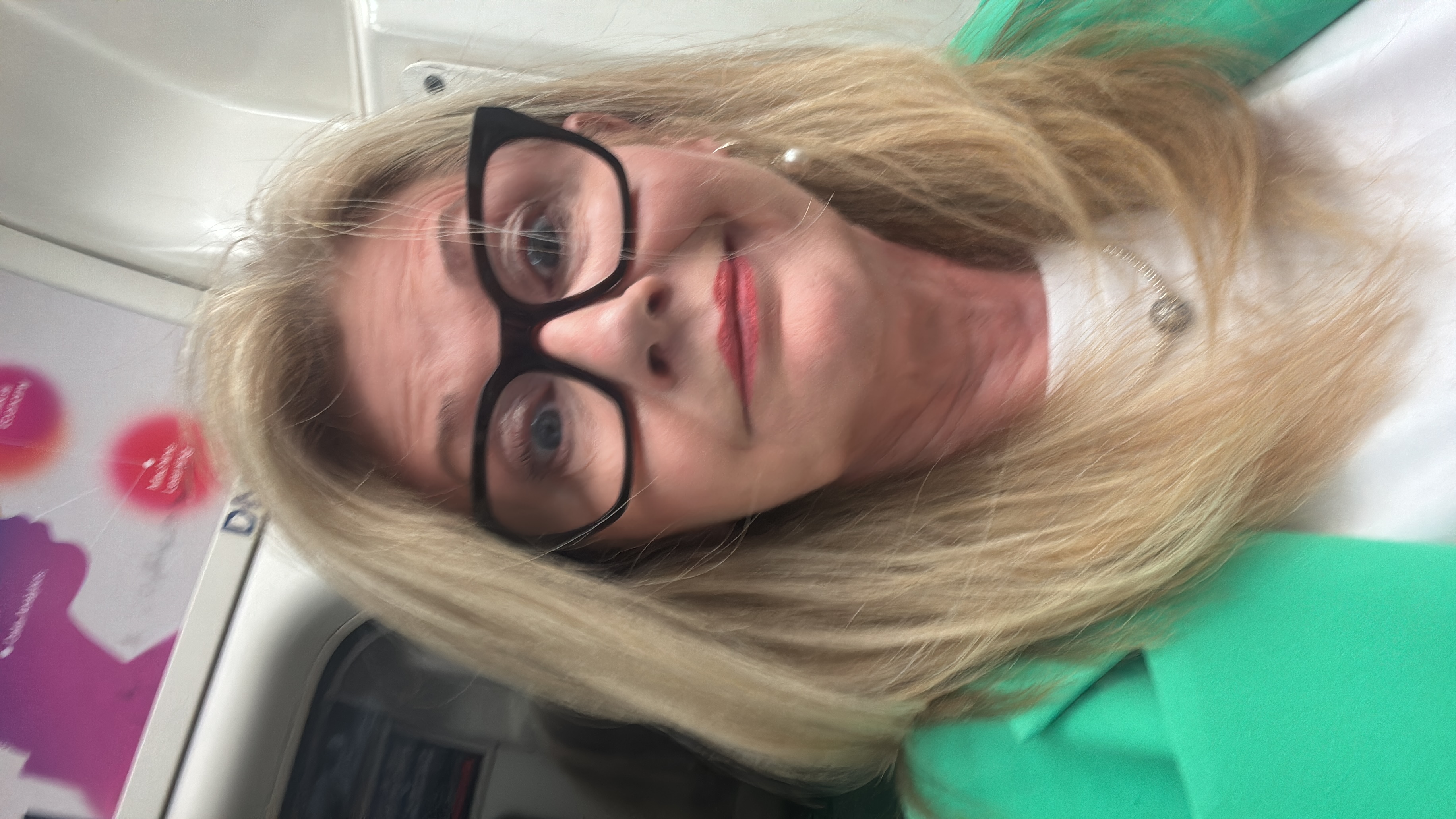 Woman with fair hair and glasses, looking annoyed. She's wearing a light top and green jacket. Appears to be sitting on the London tube.