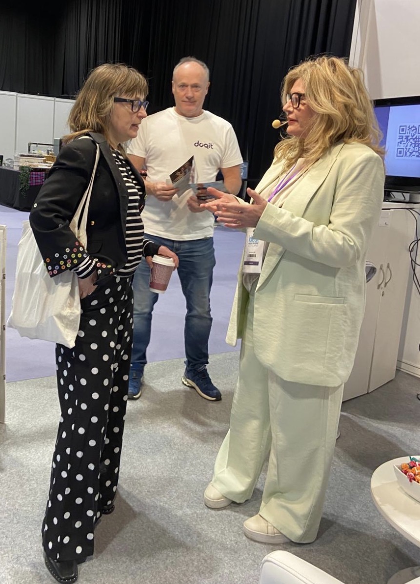 Woman in light green suit talking with another woman wearing dark jacket, striped top and dark spotty trousers - she has a light back over her shoulder. In the background is a man wearing jeans and a white t-shirt.