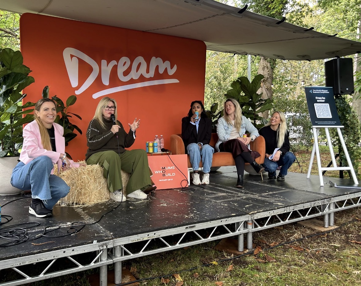 Five women sitting together on the outdoor Acorn Stage at Ideas Fest. Catherine Ann is seated on a straw bale, speaking into a microphone during the Mums Who Build panel, with a large orange Dream sign behind the group.