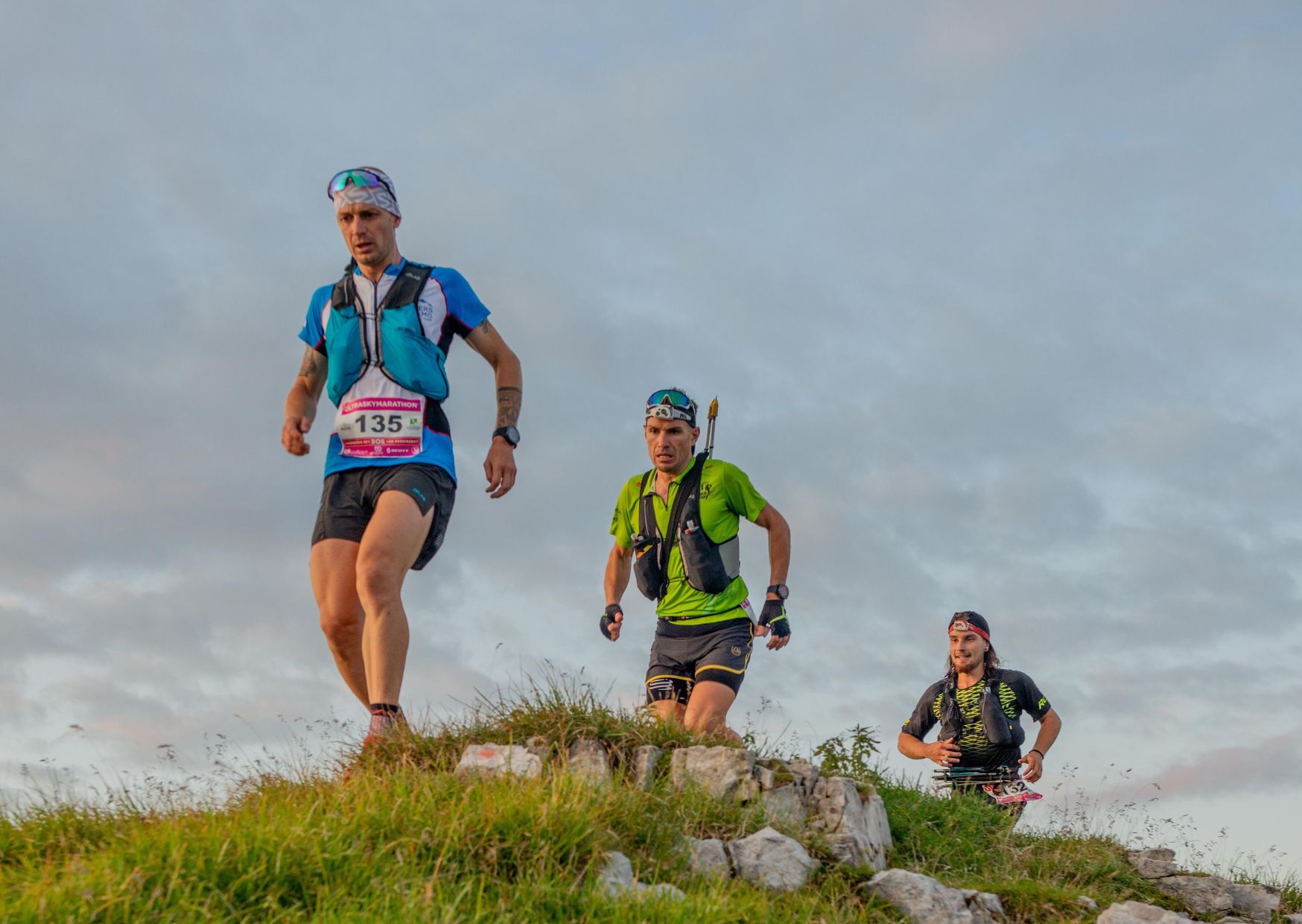 Three men all wearing running gear, carrying back packs. They all have numbers on their shirts, and appear to be at the summit of a rocky hill. The weather is grey and cloudy. 