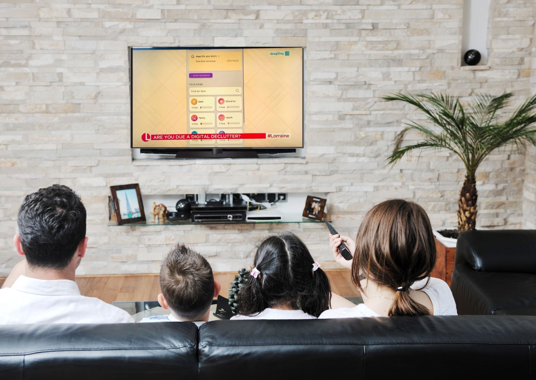 Image shows the back of heads of a family, sitting on a dark sofa. They are looking at a tv attached to a light brick wall. The image on the screen is doqit with the Lorraine logo.