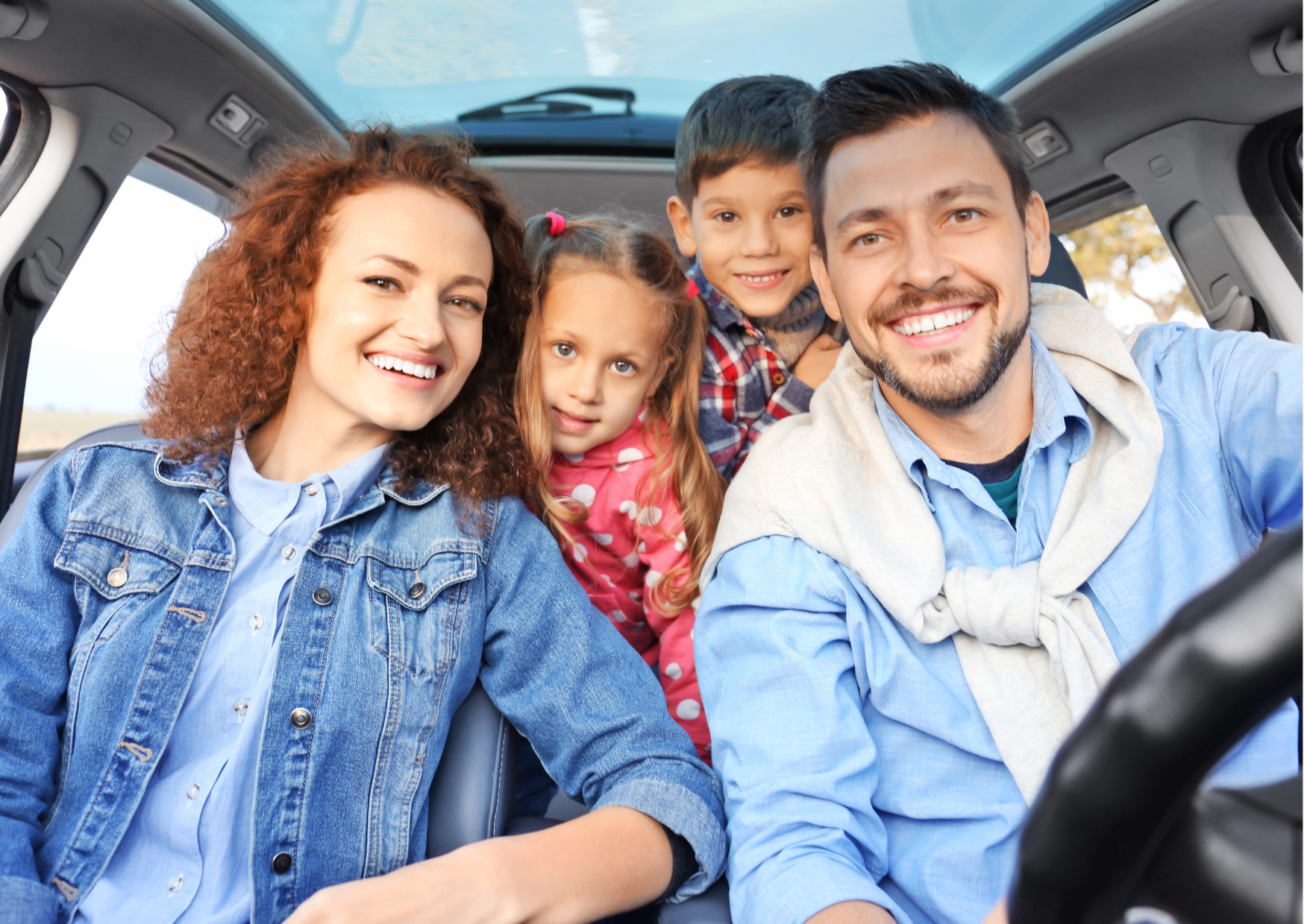 Family in a car. Male and Female in the front both wearing light blue tops, with two children in the back looking over their shoulders. The car has a glass sunroom. They all look happy.
