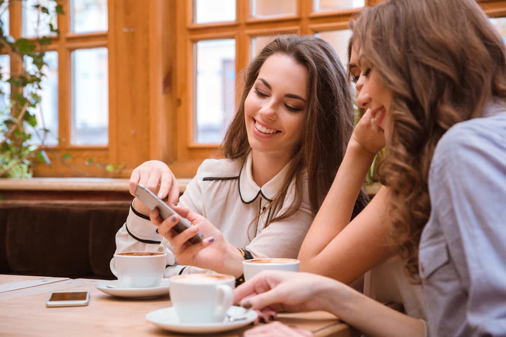 Image shows young women sitting enjoying a coffee while looking at a mobile phone.