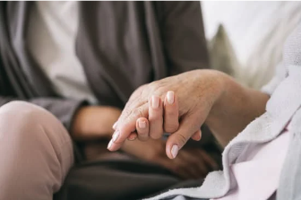 Image shows a young hand gently holding a younger hand. The younger hand is wearing brown clothing, the older appears to be in bed.