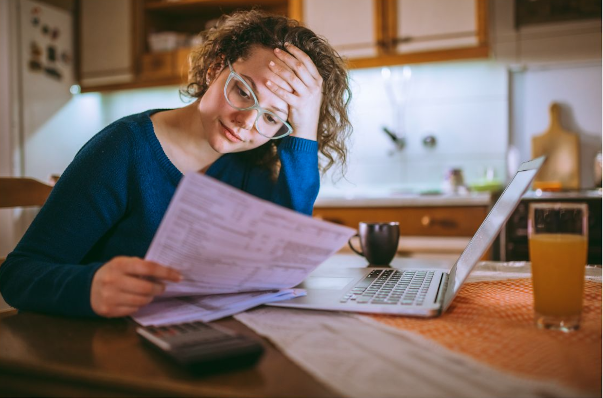 Woman at kitchen table reviewing paperwork with laptop, looking overwhelmed by household admin.