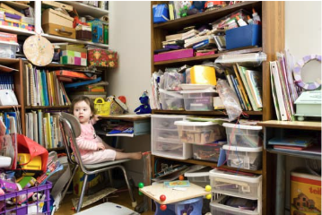 Cluttered home shelving and desk with papers, folders, and storage boxes, with a child sitting nearby — a busy, slightly chaotic everyday family space.