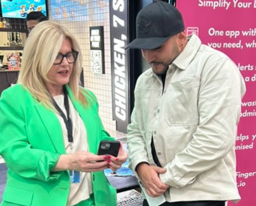 A woman in a bright green suit shows an app on her phone to a man at an exhibition booth during a live event.